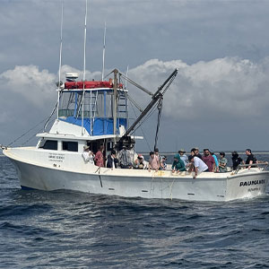 White boat sailing in the ocean with people standing and holding ropes off the back of the boat.