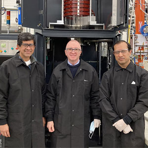 Three men in black lab coats stand in a laboratory next to a large piece of equipment, smiling and posing for the photo.