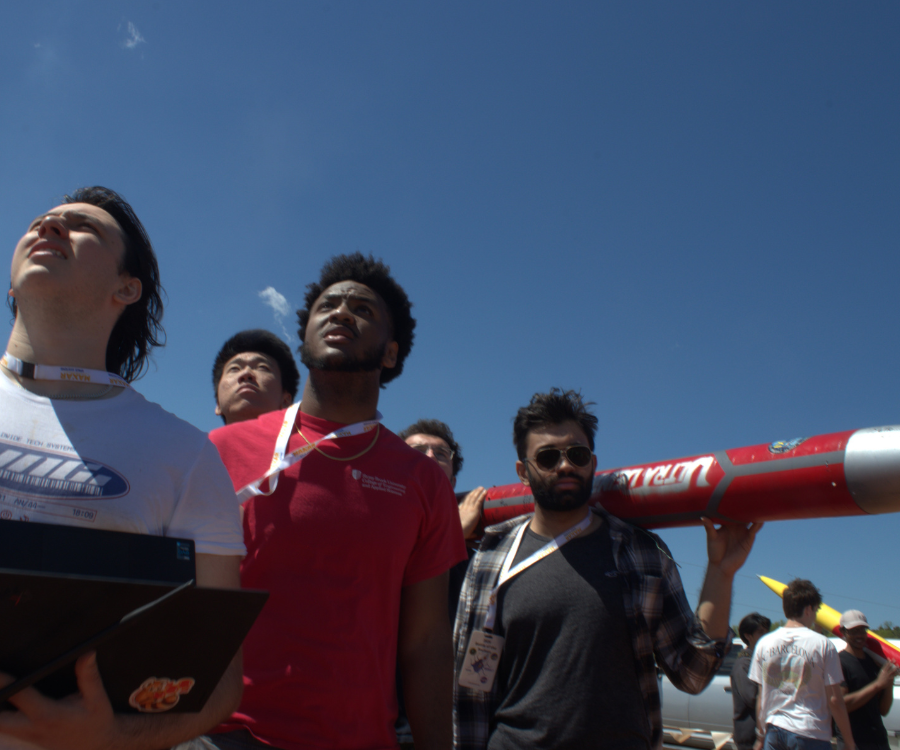 Four young men look upward while carrying a long, red cylindrical rocket body on their shoulders against a clear, bright blue sky on a sunny day.