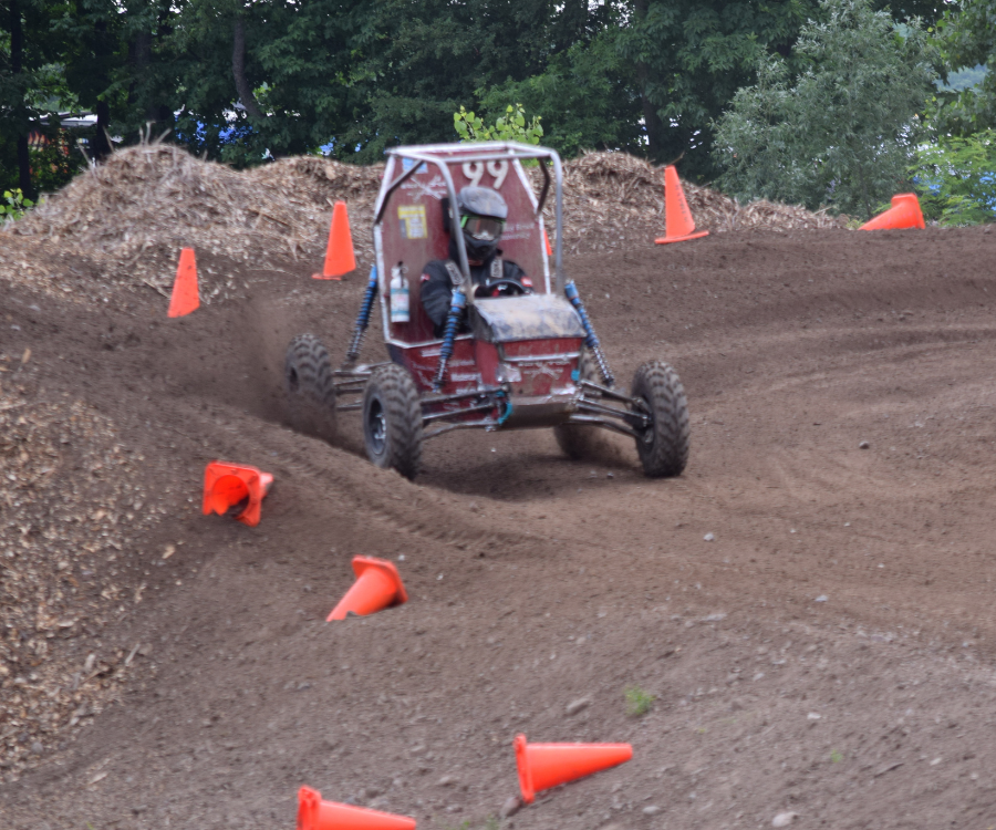 A driver in a helmet operates a red Baja SAE race car with the number 99 on a dirt track marked by orange traffic cones near a pile of wood chips.