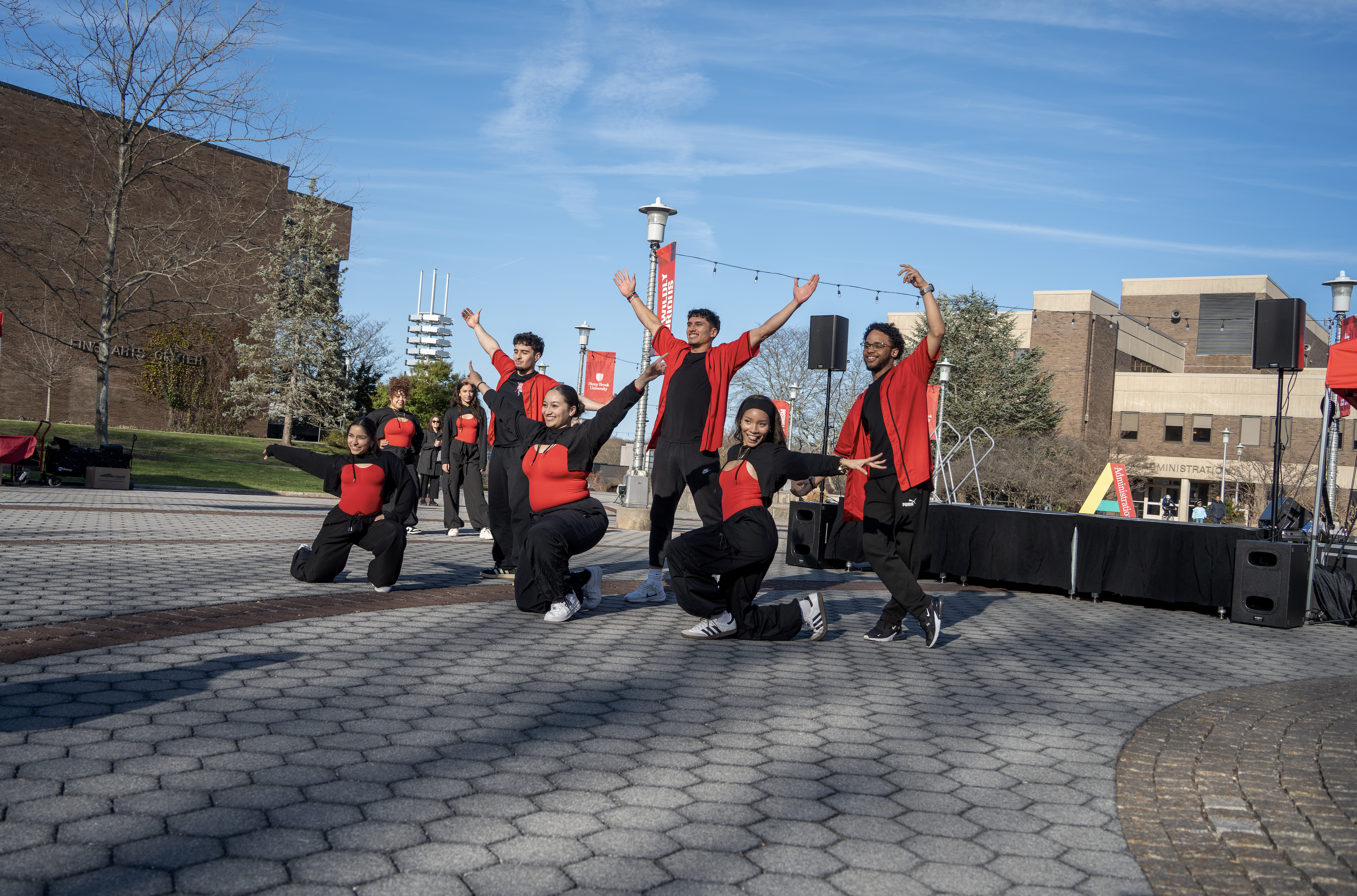A group of student dancers in matching red and black outfits performing a routine on a paved campus plaza under a blue sky.