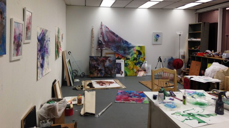 A photo of a graduate student's studio space, cluttered with various paints, fabrics, and canvases.