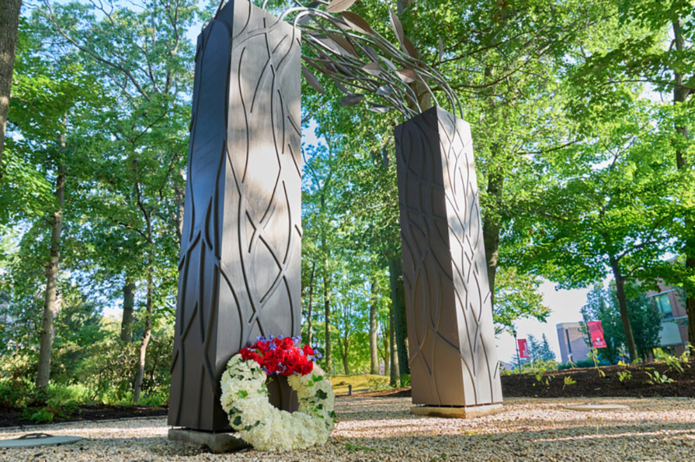 Metal archway sculpture in a wooded area with a floral wreath at its base.