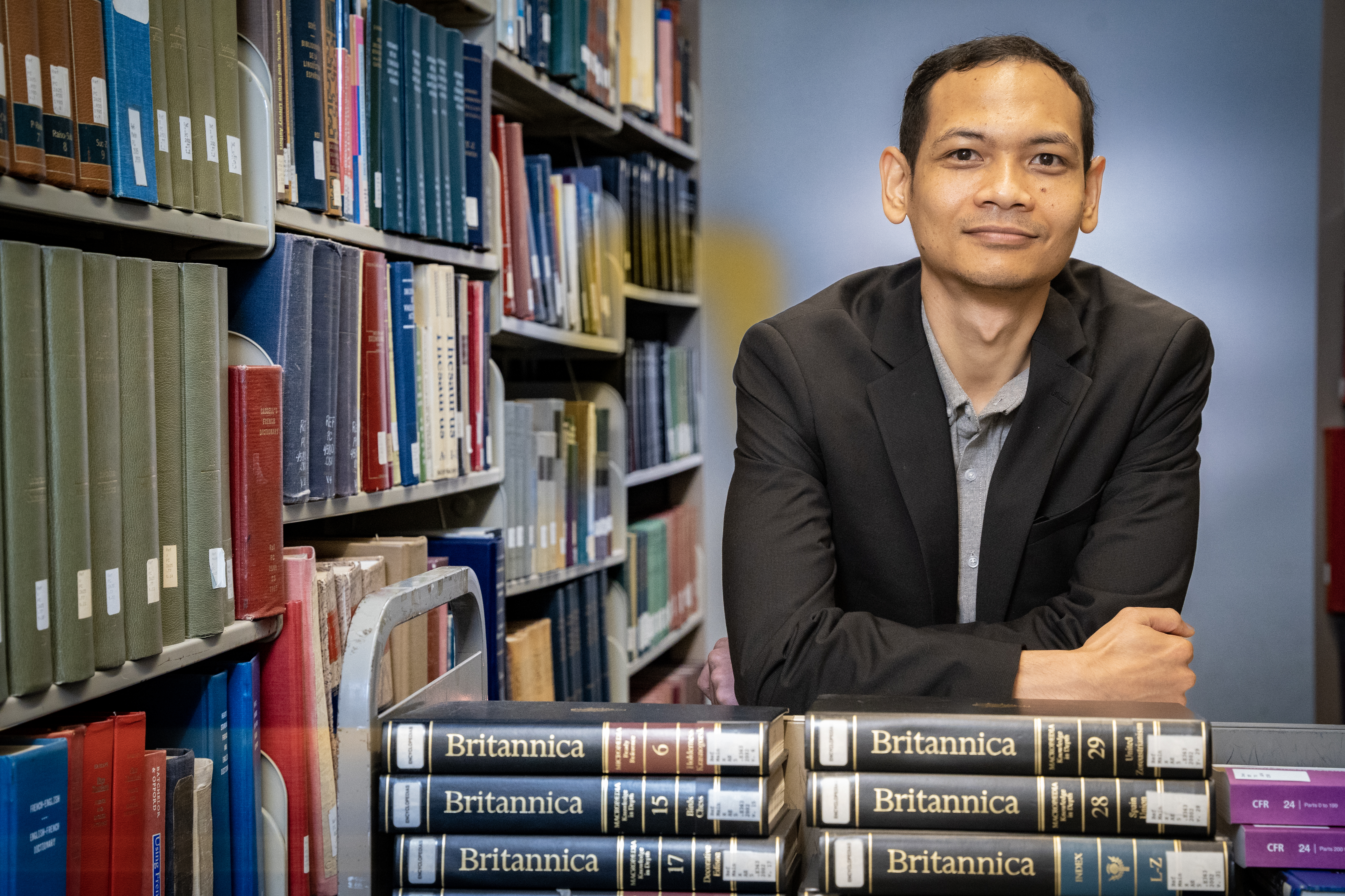 Southeast Asian man wearing black blazer, leaning on books on a library cart and standing in front of shelf of books