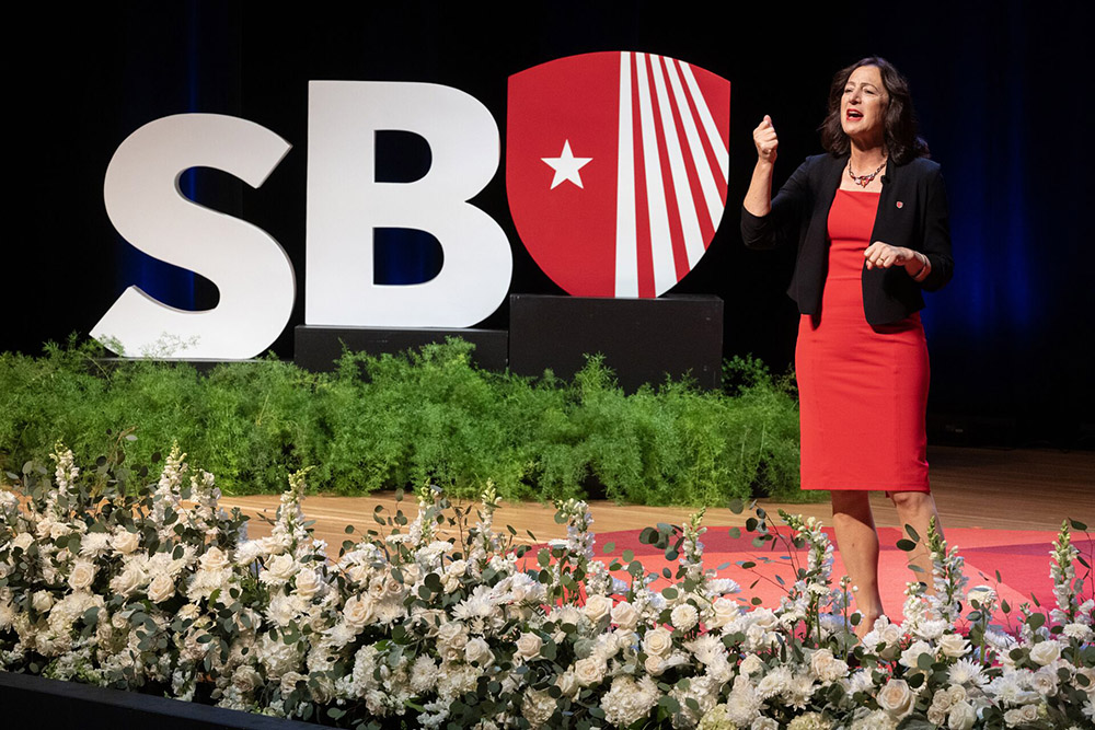 President Goldsmith in a red dress and black blazer speaks on a stage with a large "SBU" logo background, accented by a floral display at the front.
