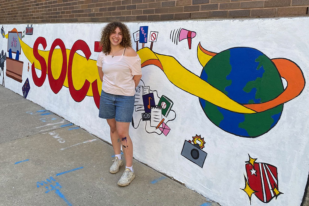 student standing infront of SOCJ mural with earth and other sympbols on it