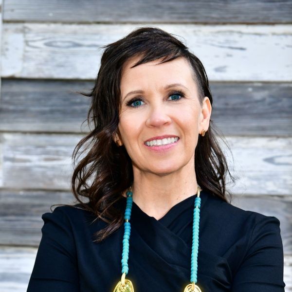 Dr. Nichole s. Prescott Person wearing a black top and a large gold necklace, smiling in front of a wooden plank background.
