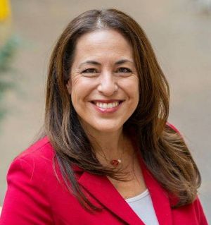 AnnaMaria Gounaris Smiling woman with long brown hair in a red blazer and light top, standing outdoors. The background is softly blurred, creating a warm, approachable tone.