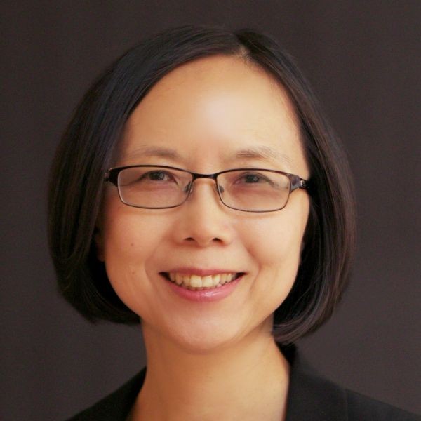Dr. Amy Liao Portrait of a smiling person with glasses, wearing a black blazer over a gray top, standing in front of a dark background.