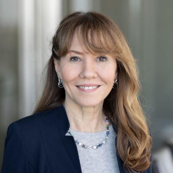 Maria Pasquale Professional portrait of a smiling individual with long brown hair, wearing a dark blazer and a silver necklace.