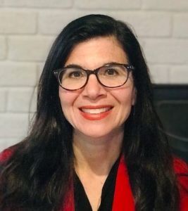 Sheri Levy Portrait of a smiling person with glasses, wearing a blue top, set against a neutral background.