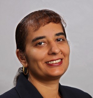 Surita Bhatia A woman in a black blazer smiles confidently against a gray background. She has medium-length hair, hoop earrings, and wears light makeup.