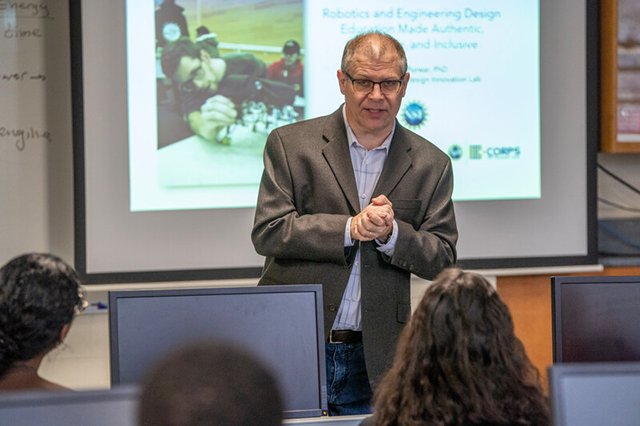 Mechanical Engineering Faculty Professor Jon Longtin in front of his classroom with a slide behind him of a mechanical engineer at work