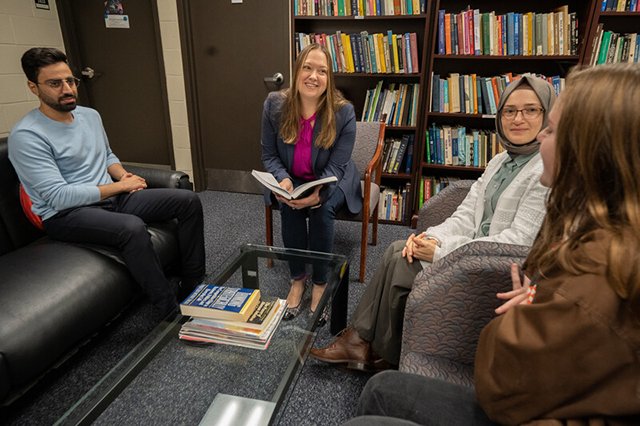 Technology and Society Faculty Professor Elizabeth Hewitt is seated in a library and surrounded by four students, and she is smiling with a book in her hand