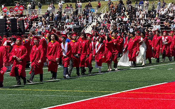 Graduates in red caps and gowns walk across a green football field, with an audience in the background.