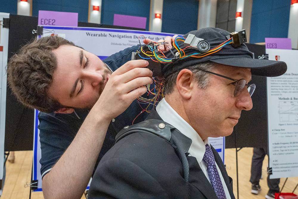 Dean Andrew Singer at Engineering Senior Design Showcase A man adjusts colorful wires on another man's cap-mounted tech device at a science exhibit. They collaborate closely, focused on innovation.