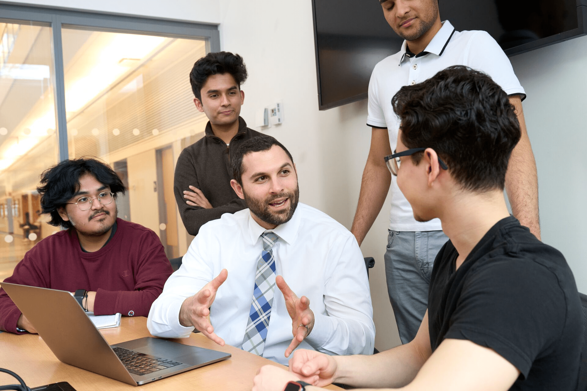 A group of five people, engaged in a friendly conversation around a table. A man in a white shirt gestures while others listen attentively. A laptop is open.