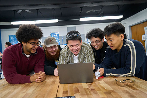 A diverse group of five young adults enthusiastically gathers around a laptop on a wooden table, sharing a moment of excitement in a brightly lit room.