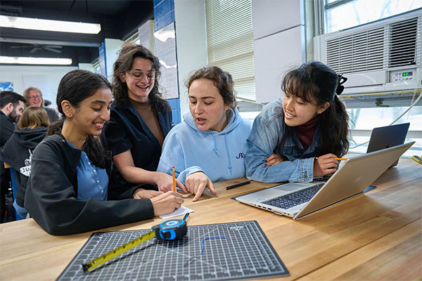 Four diverse young women collaboratively engage in a creative task at a wooden table, surrounded by school supplies and a laptop.