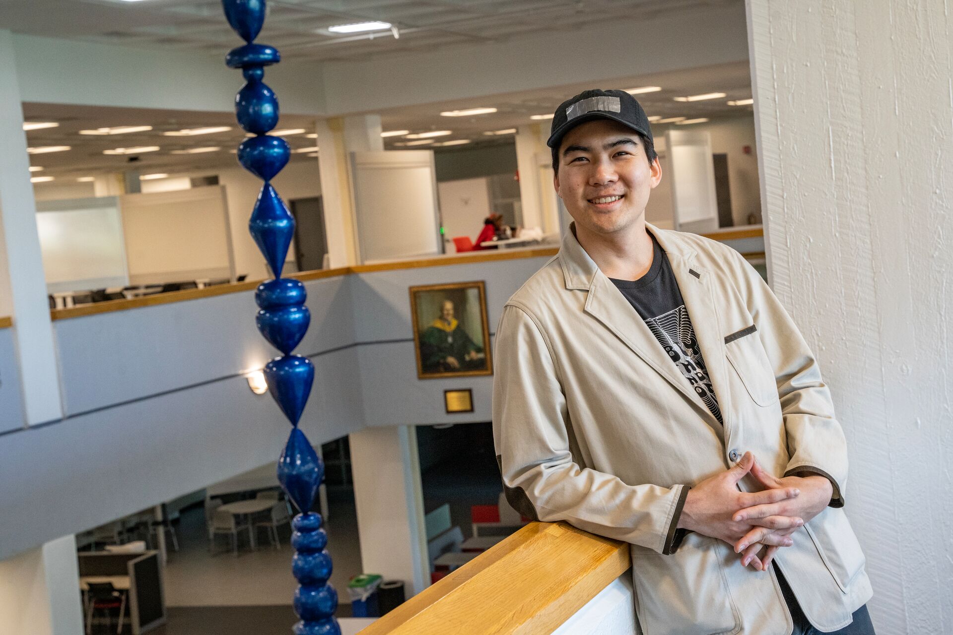 image of sy lee smiling while leaning on a 2nd floor ledge in the library at stony brook university where his water droplet sculpture hangs from the ceiling in the background