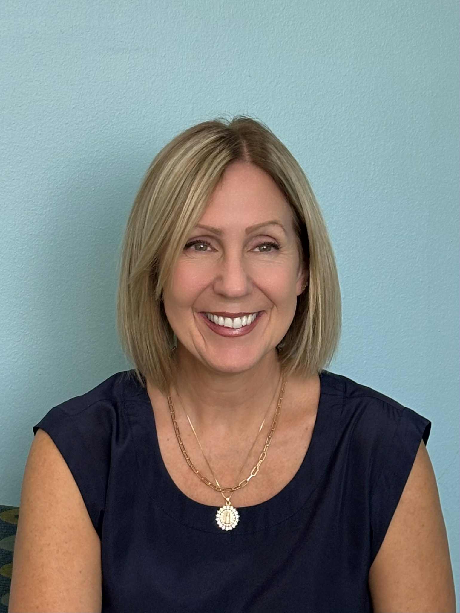 A woman with blonde hair wearing a necklace sits comfortably on a couch, looking relaxed and engaged.