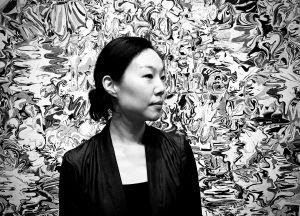 Sohl Lee A person stands calmly against a swirling, abstract black-and-white backdrop. The scene conveys a sense of contemplation and artistic intrigue.
