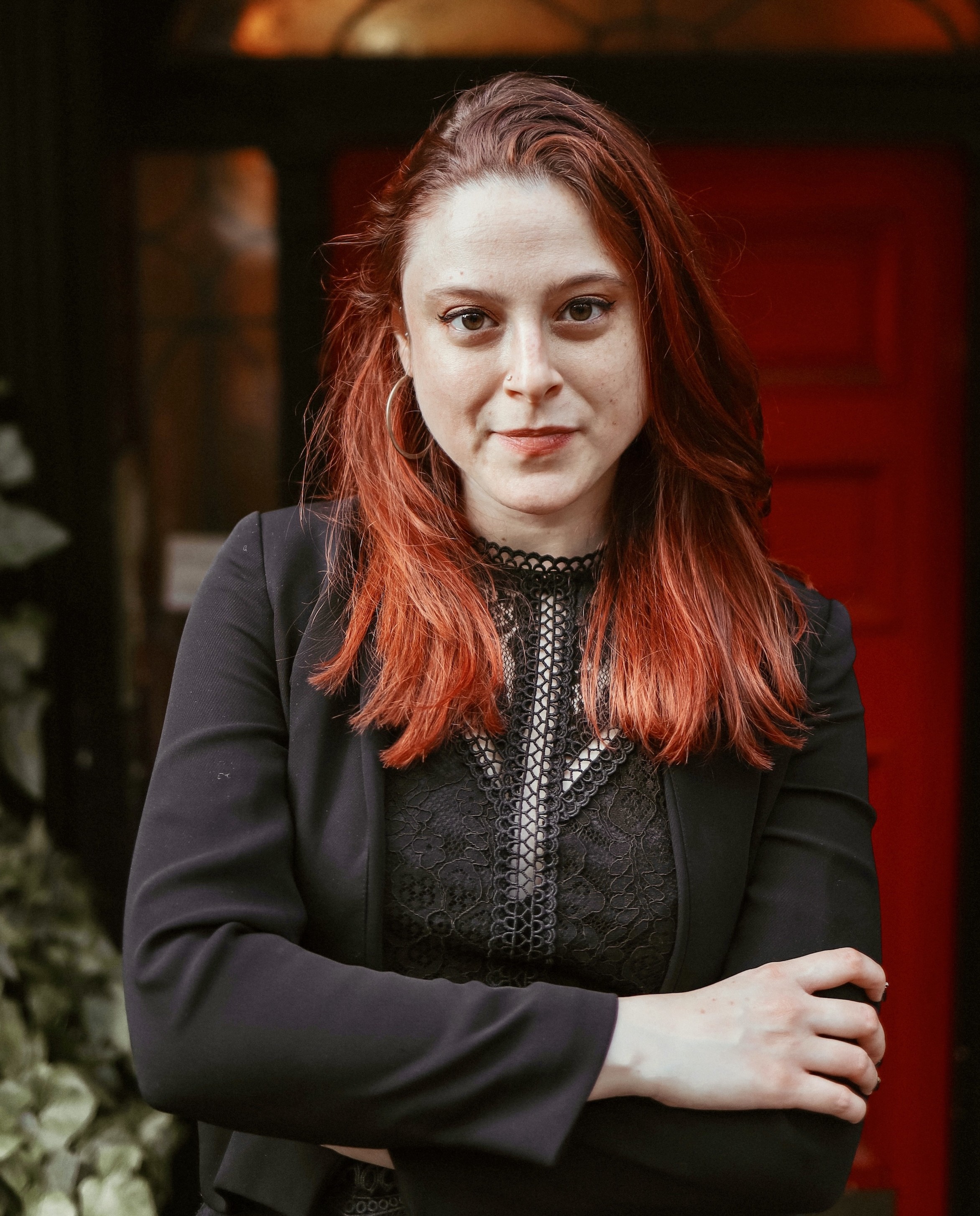 a woman with a slight smile stands, arms folded, in front of a reddish doorway next to a green plant