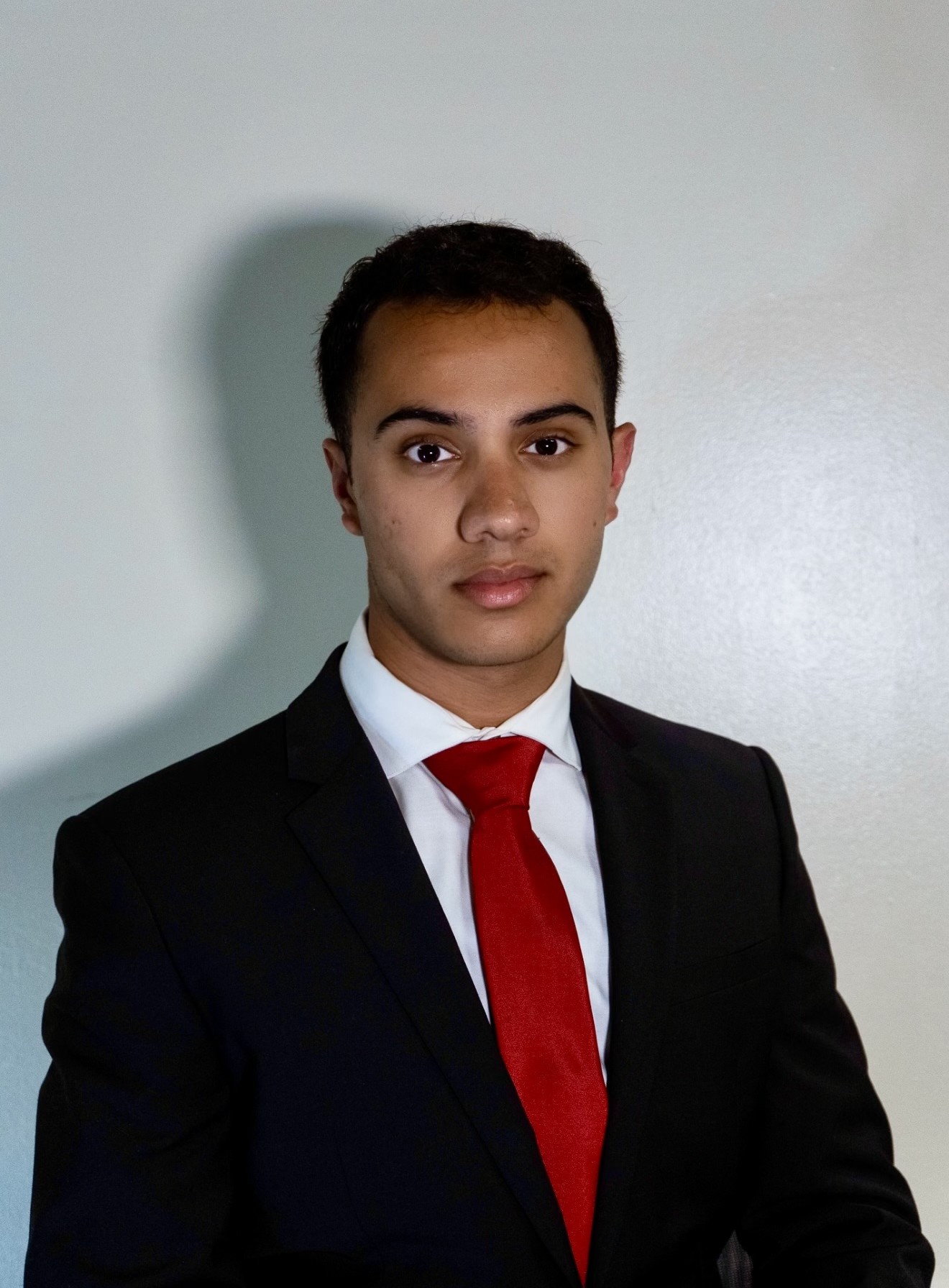 Portrait of a person wearing a black suit and red tie, standing against a light-colored background.