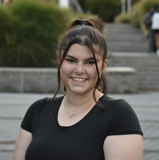 Portrait of a smiling person with pulled back hair, wearing a black top and a necklace, outdoors.