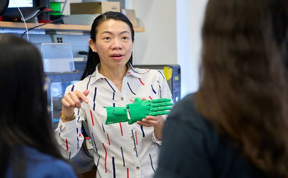 A person in a patterned shirt holds up a green prosthetic hand, engaging in discussion with two others. The setting is a laboratory, conveying learning and innovation.