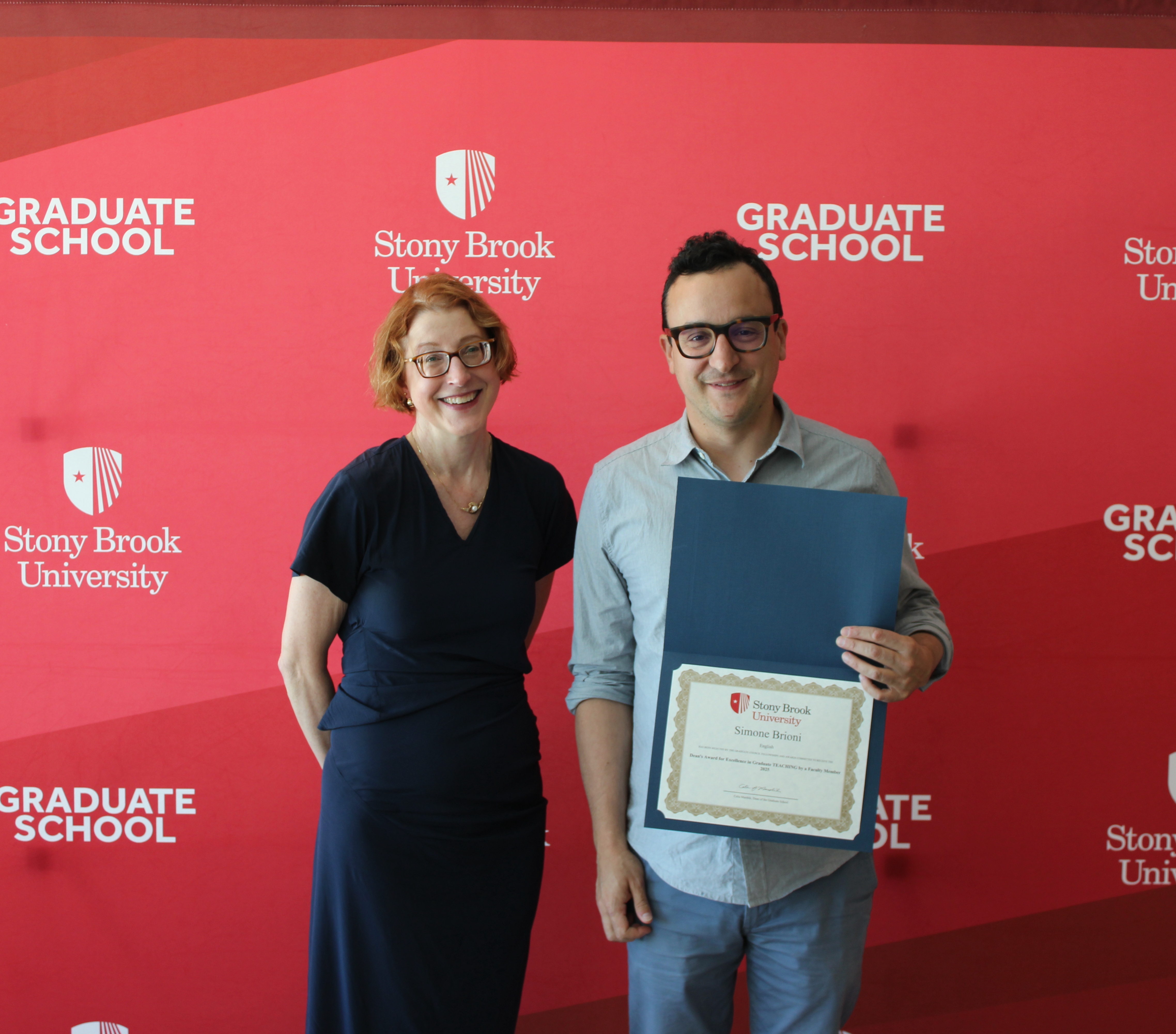photo of Simone Brioni, award winner, and Celia Marshik, Dean of the Graduate School standing next to each other. Simone is holding his award for graduate teaching, and they're in front of a red background