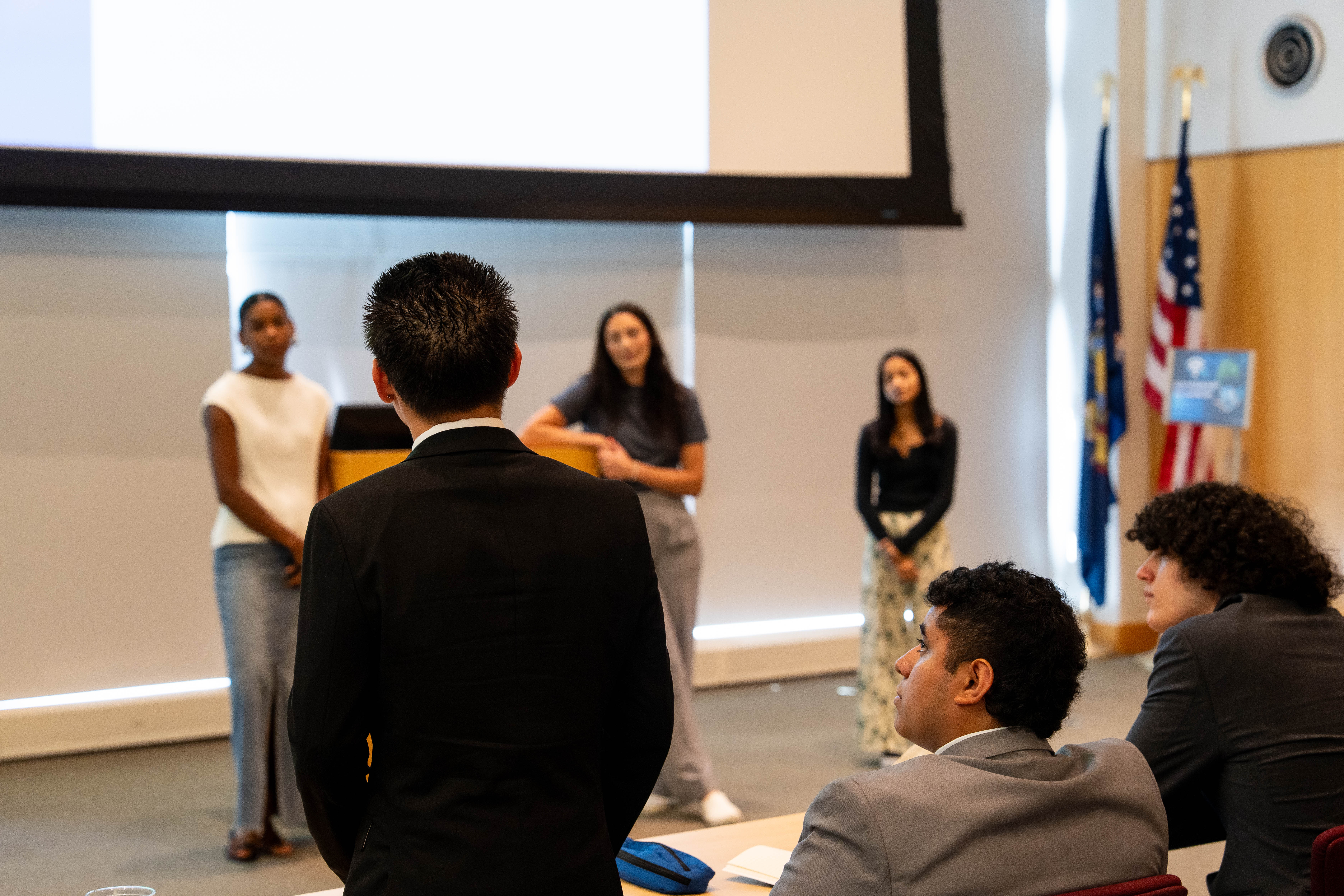A man in a black suit stands with his back to the camera, facing three women who are standing at the front of a room, possibly during a presentation or Q&A session. The three women are standing in front of a large, blank projection screen; one wears a white sleeveless top and long denim skirt, the middle wears a grey t-shirt and light trousers, and the one on the right wears a black long-sleeved top and patterned pants. In the foreground, two other men are seated at a table, looking up toward the presenters. The background includes wood-paneled walls, an American flag, and a New York State flag.