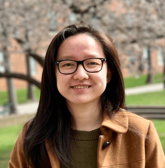 A professional headshot of a smiling young woman with long dark hair, wearing black-rimmed glasses, an olive green sweater, and a tan coat. She is standing outdoors on a campus, with a blurred brick building and blooming cherry blossom trees in the background.