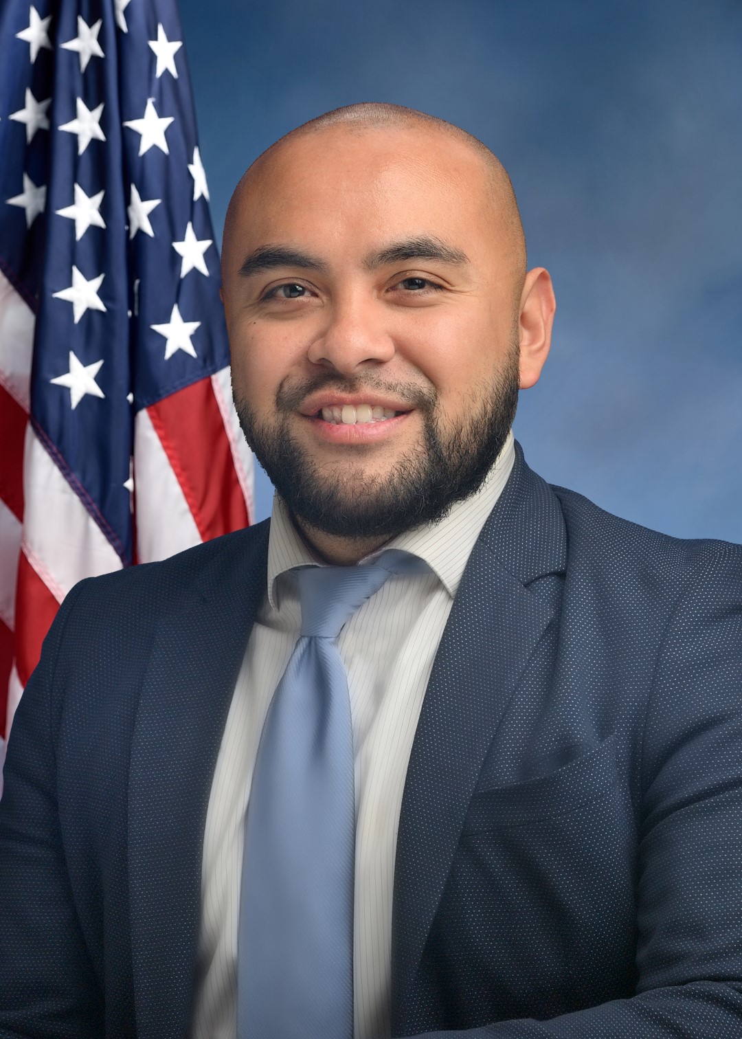 An individual dressed formally in a blue suit, white shirt, and light blue tie stands in front of a backdrop featuring the American flag and a gradient blue background. 