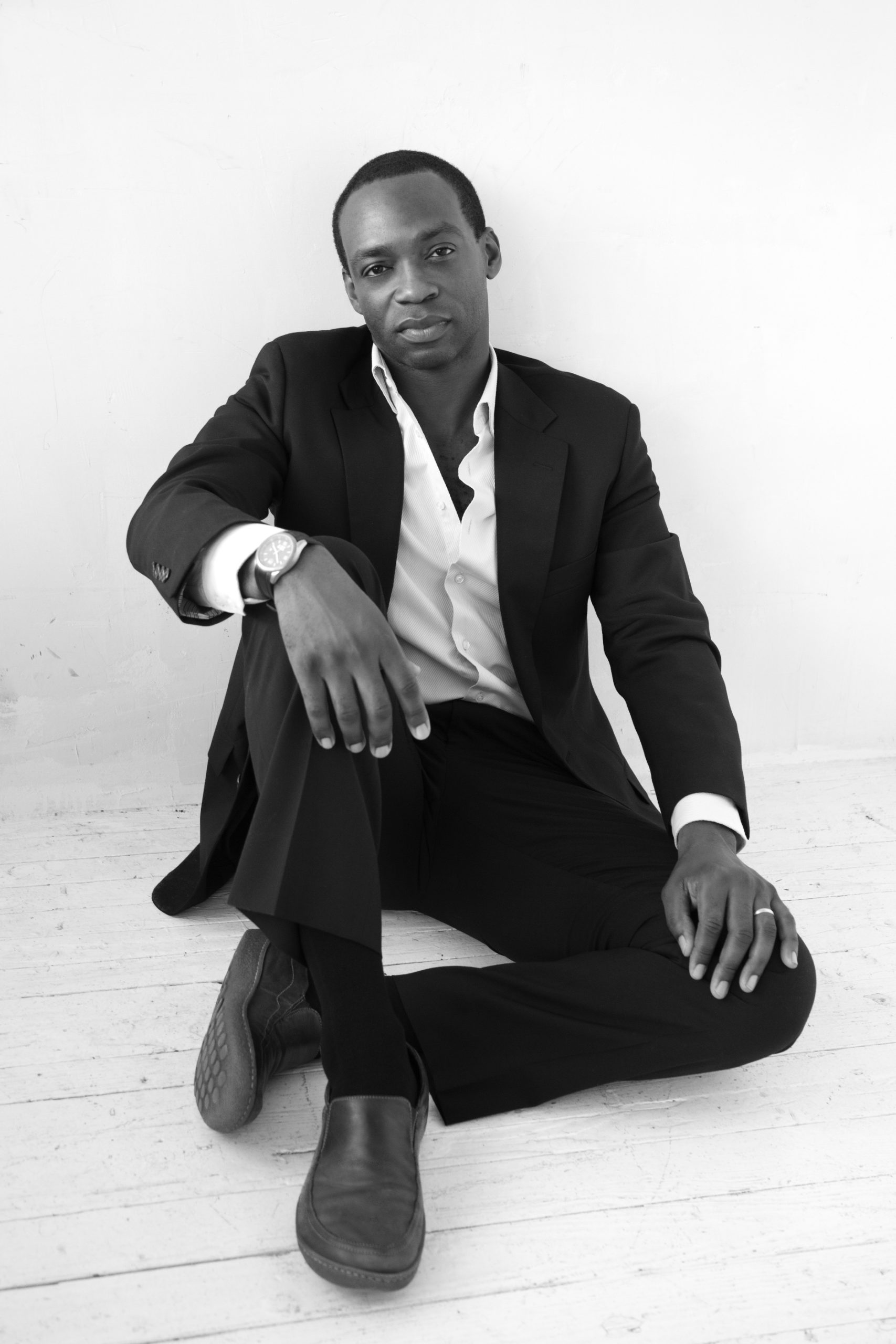 man lounges on the floor leaning against the wall with his hands on his knees whearing a suit with no tie