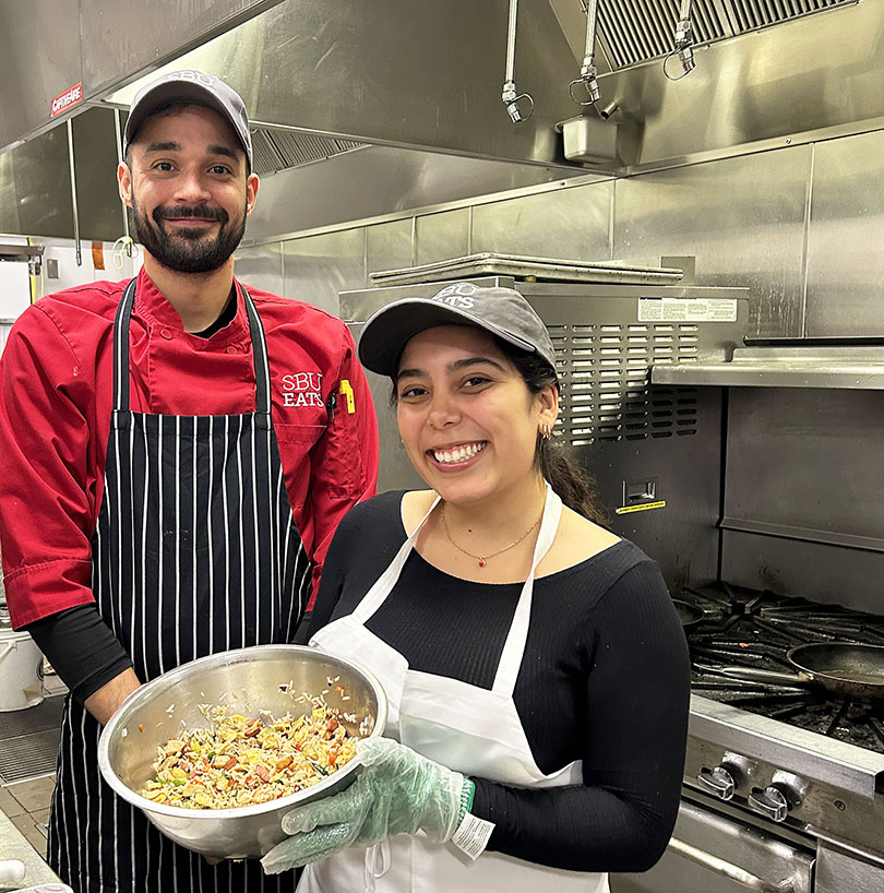 Two chefs in a commercial kitchen smiling while holding a large mixing bowl of fried rice.