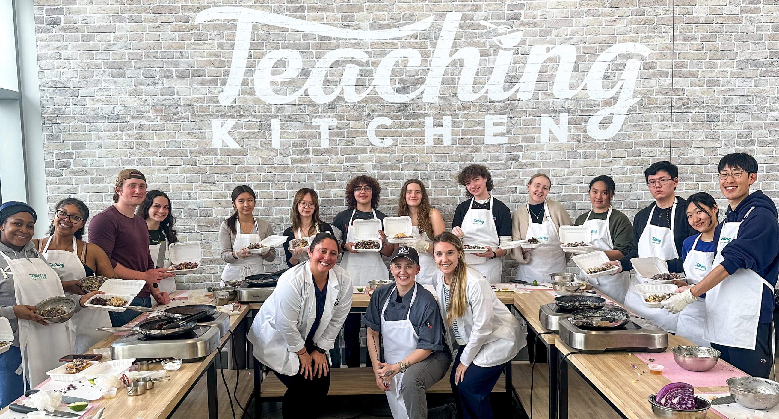 Group photo of a diverse cooking class in a kitchen, with participants holding food containers and wearing aprons, against a backdrop that says "Teaching Kitchen."