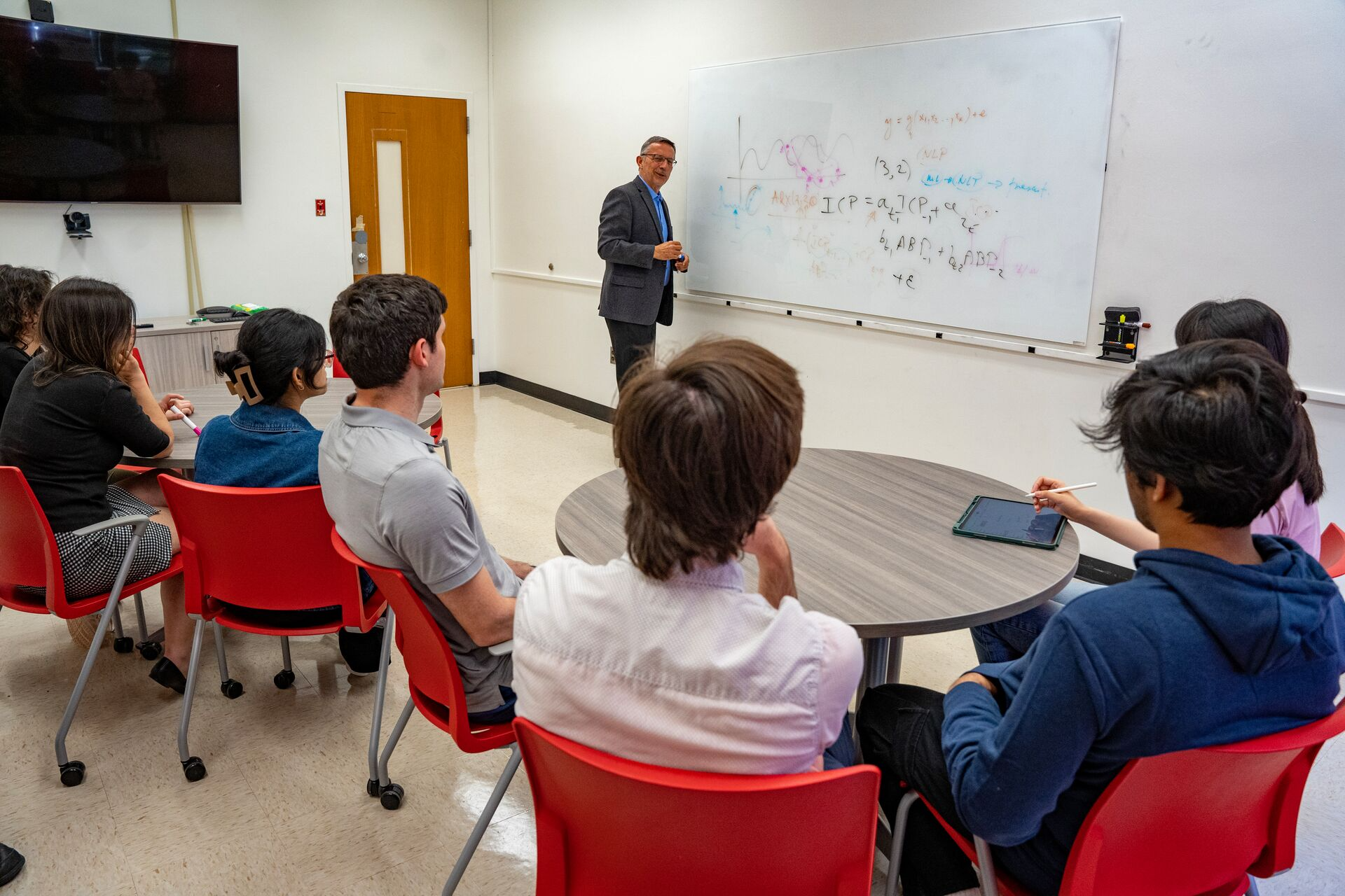 A teacher stands at a whiteboard covered in diagrams, instructing a group of attentive students seated around a table in a classroom setting.