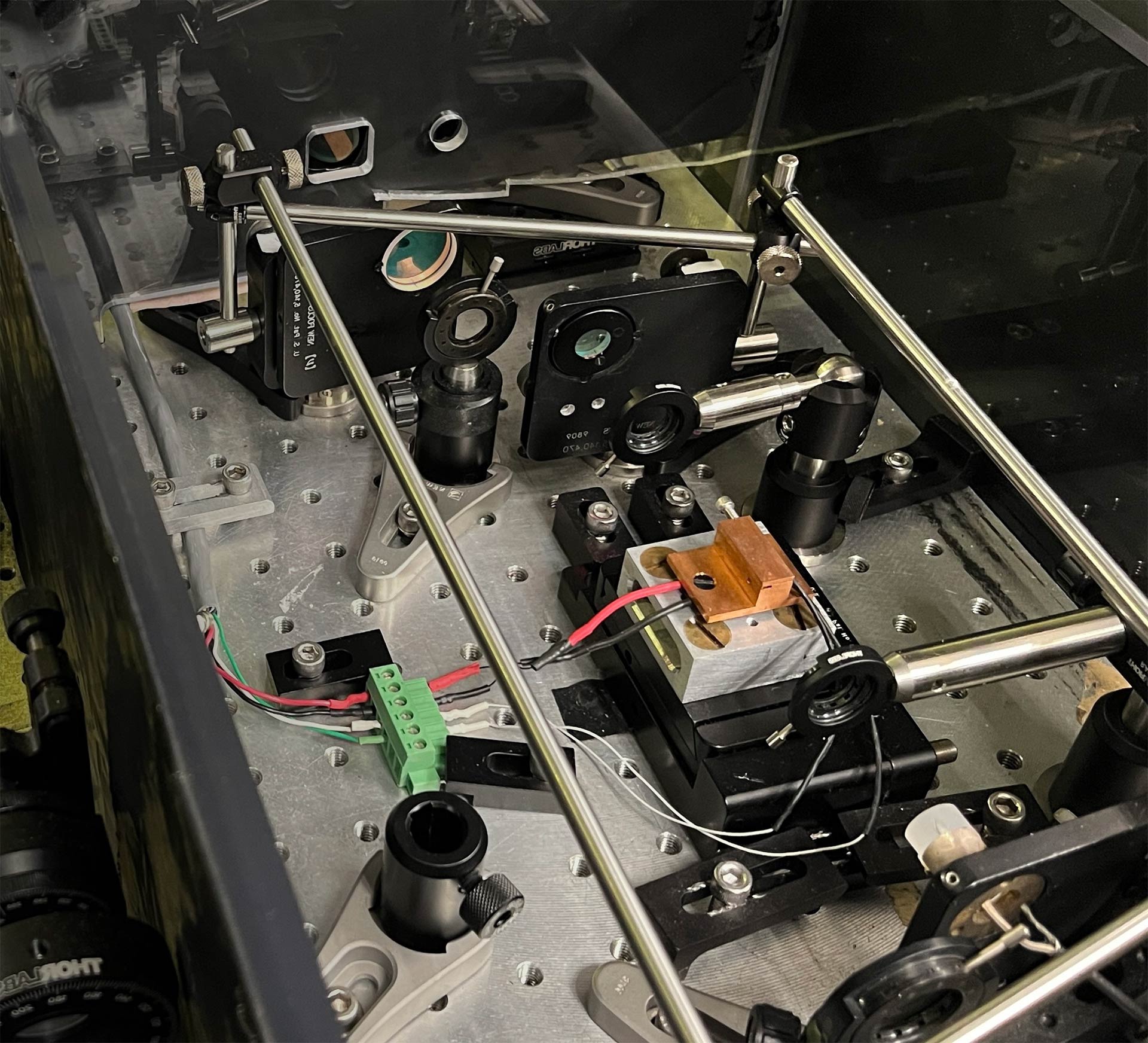 An optical breadboard setup with mirrors, lenses, and lasers mounted on a metallic grid table.