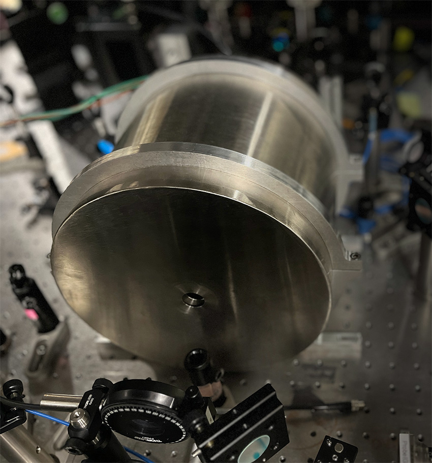 A large cylindrical metallic component mounted on an optical breadboard table inside a laboratory.