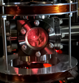 A metallic vacuum chamber with a circular viewport reveals a glowing pinkish-red interior, sandwiched between large copper wire coils.