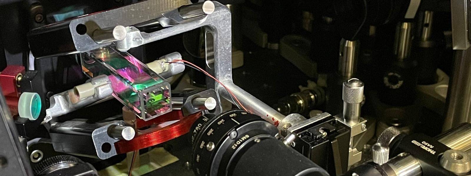 A rectangular glass cell glowing with pink and green light is mounted inside a complex arrangement of metallic laboratory optics and adjustment mounts.
