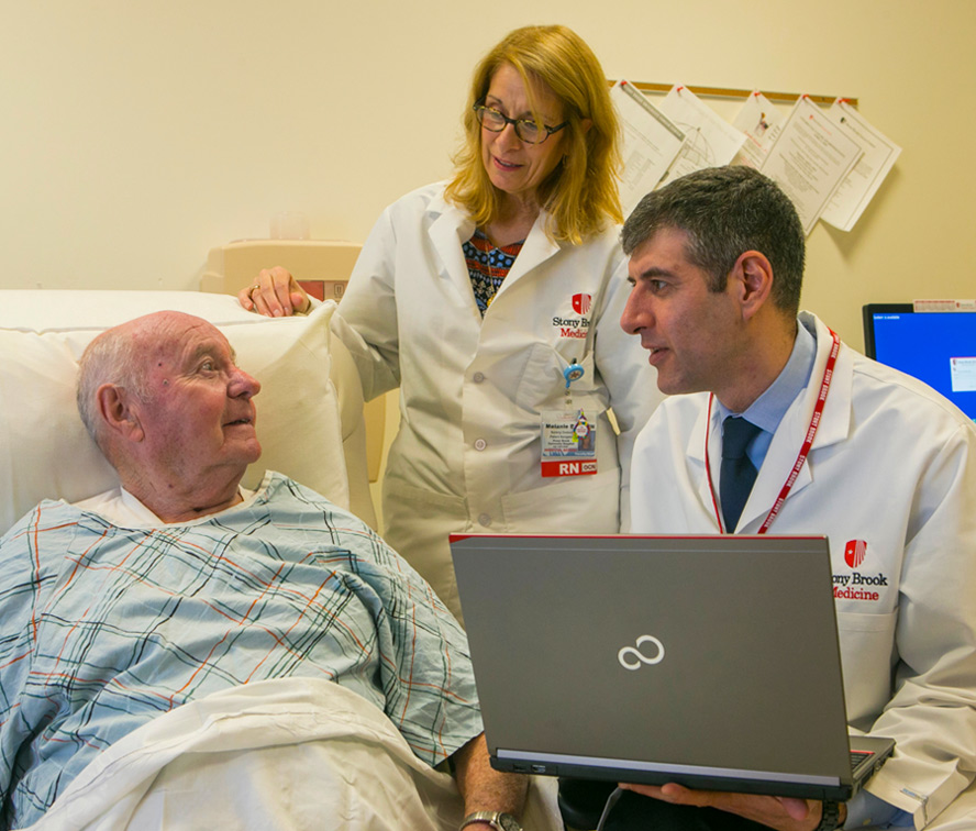 two doctors sitting bedside with an elderly patient, one doctor is holding a laptop computer