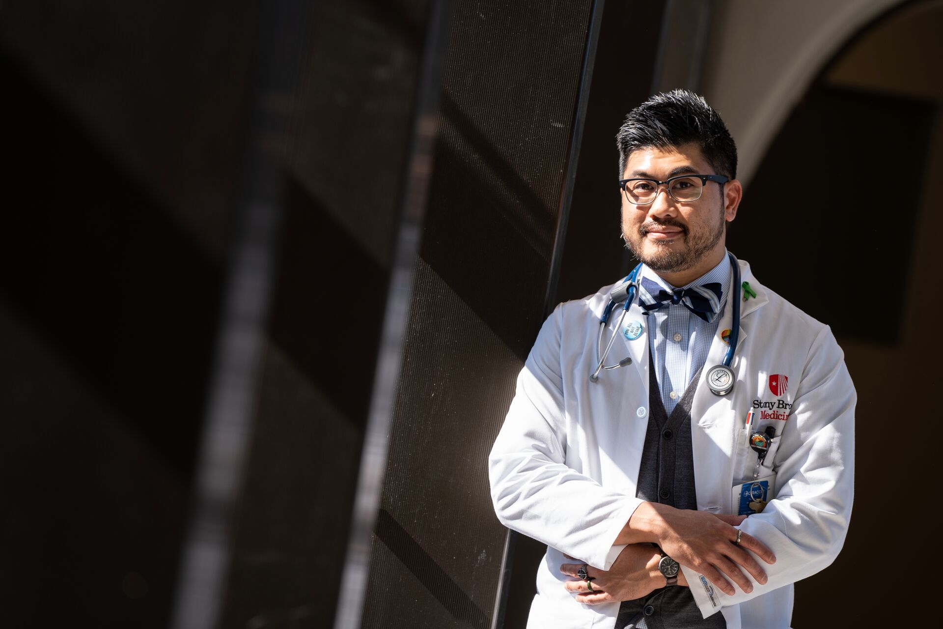 A male healthcare professional wearing a white lab coat, stethoscope, and bowtie poses with arms crossed.