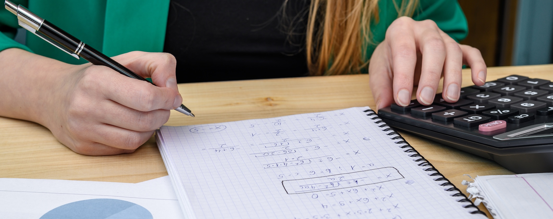 Person writing math equations in a spiral notebook next to a desktop calculator
