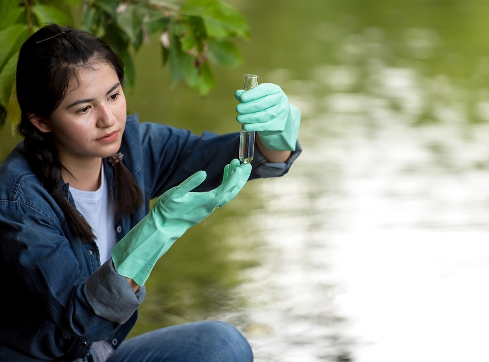 A young researcher wearing green rubber gloves inspecting a clear water sample in a test tube outdoors near a lake.