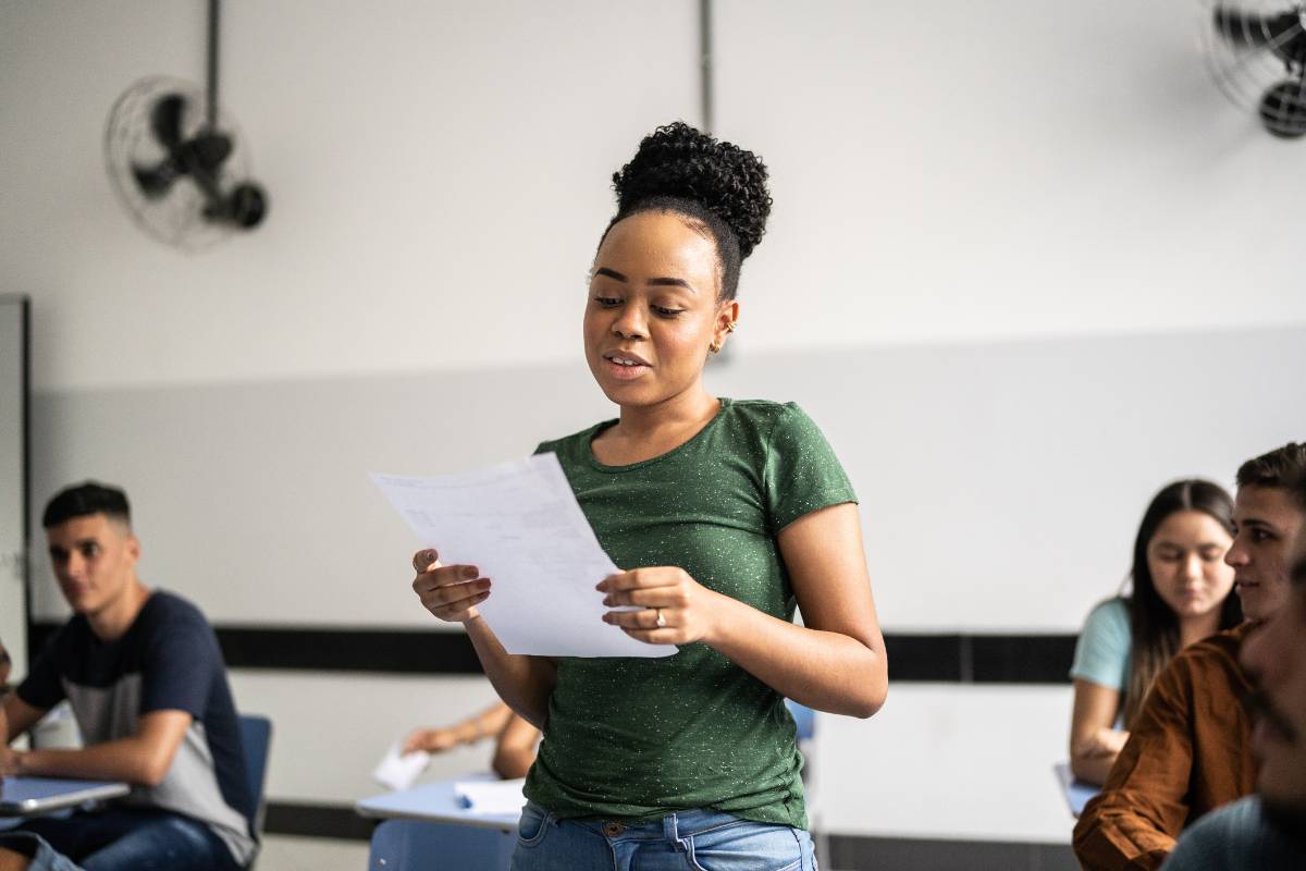 A young woman standing at the front of a classroom reading aloud from a sheet of paper to her seated classmates.