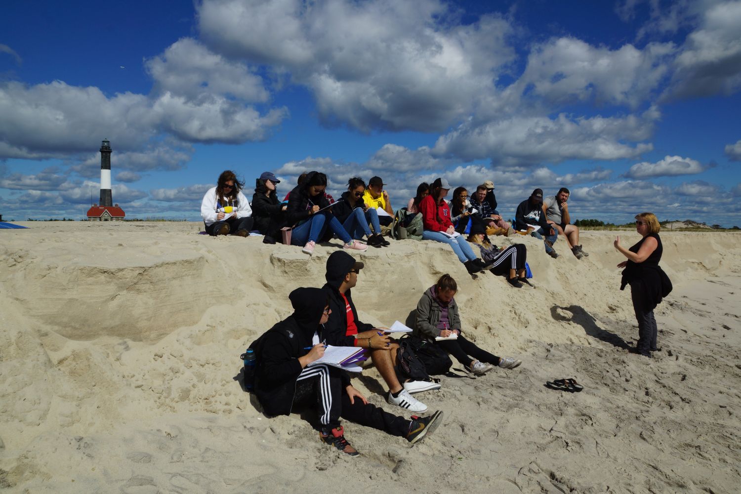 Students sit on a jetty at a beach listening to their professor
